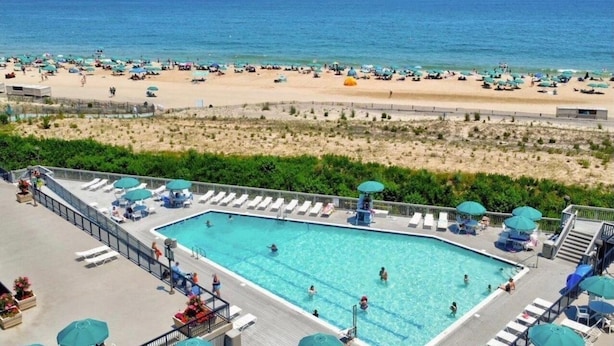 A pool area with lounge chairs and umbrellas, a beach in the background, and a clear view of the ocean. - Breathtaking Ocean View, Beachfront Condo-Steps to Beach/Pools, Sea Colony East (Bethany Beach)