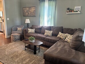 A living room with a brown sectional sofa, a small round ottoman, a coffee table with a plant, a world map on the wall, and a shelf with plants and a sign.