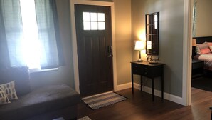 A room with a dark wooden door, a small table with a lamp, a sofa, and a bed in the adjacent room. - Cozy house on a quiet street (Bedford)