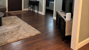 A living room with a ceiling fan, a dark wooden door, a lamp, a framed picture, a television, and a doorway leading to another room. - Cozy house on a quiet street (Bedford)
