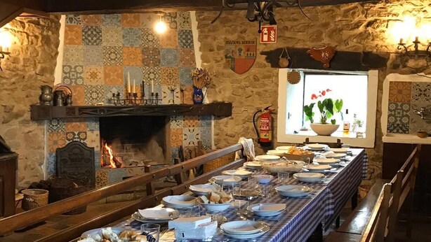 A long dining table set with plates, glasses, and cutlery, a fireplace, and a stone wall with a tiled backsplash.