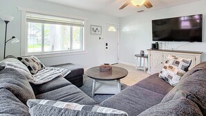Ein modernes Wohnzimmer mit Deckenventilator, Flachbildfernseher, Sofagarnitur, Couchtisch und einem Fenster mit Jalousien.