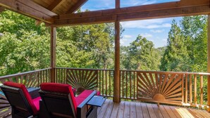 A wooden deck with a covered area, furnished with wicker chairs and a table, overlooking a forested area.