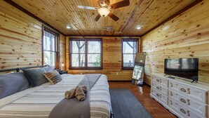 A wooden cabin bedroom with a bed, dresser, TV, and large windows.