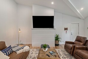 Una sala de estar con un televisor de pantalla plana montado en una pared blanca, un sofá de cuero marrón, una mesa de centro con un libro y un control remoto, y una alfombra estampada.