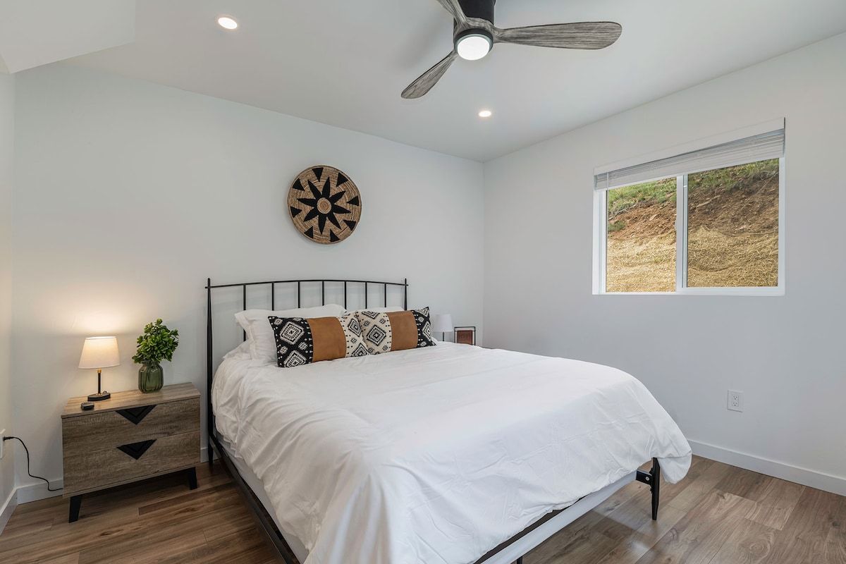 A bedroom with a bed, a nightstand, a wall-mounted decorative item, a ceiling fan, and a window with a view of the outdoors.