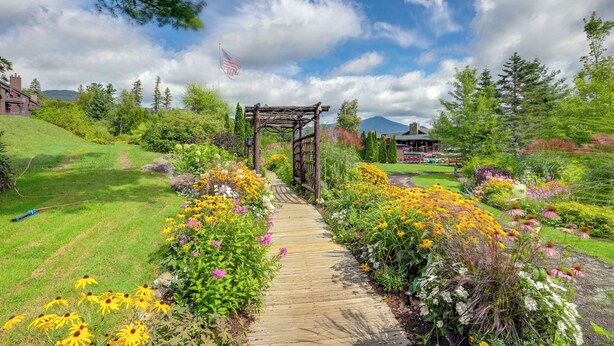 Un vialetto fiorito conduce a un gazebo di legno con una bandiera americana, circondato da fiori colorati e vegetazione.