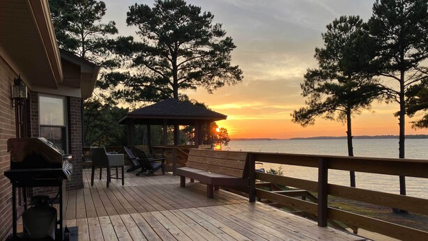 A wooden deck with a grill, chairs, and a gazebo overlooking a body of water at sunset.