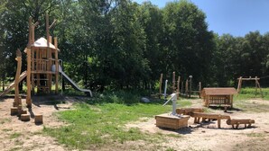 Un parc infantil amb una estructura de joc de fusta, tobogan i zona de sorra amb bancs de fusta.
