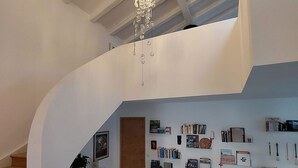 A spacious loft with a high ceiling, a modern chandelier, a staircase, and a wall adorned with various items.