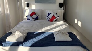 A neatly made bed with a blue blanket and two pillows, a white wall with a painting, and a window with curtains.
