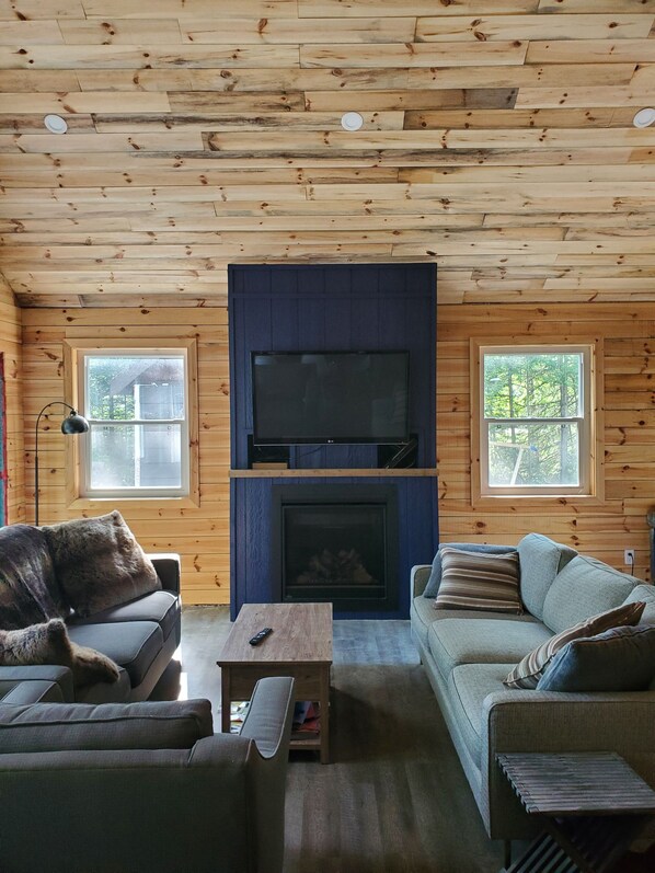 A cozy cabin-style room with a fireplace, two sofas, a coffee table, and a television mounted above the fireplace.
