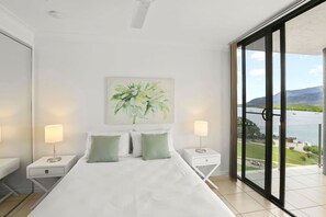 Une chambre à coucher avec un lit, deux tables de chevet, un tableau et une vue sur une étendue d’eau, visible à travers de grandes fenêtres.