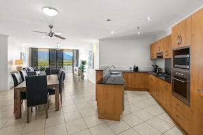 A modern kitchen with wooden cabinets, a central island, and a dining area with a view. - Seascape: Luxury Waterfront Living in the Heart of Cairns (Cairns City)