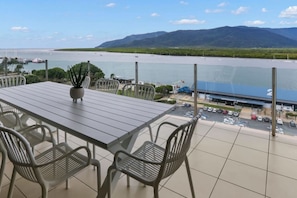 Un balcón con vista al agua y montañas, una mesa con sillas y una planta en maceta.