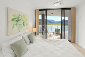 Habitación de hotel con cama, un cuadro, un balcón con vista a montañas y agua, y una silla.