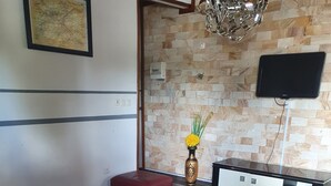 Uma sala de estar com um sofá vermelho, uma mesa de centro branca, um tapete estampado, um vaso decorativo e uma TV de tela plana fixada em uma parede de pedra.