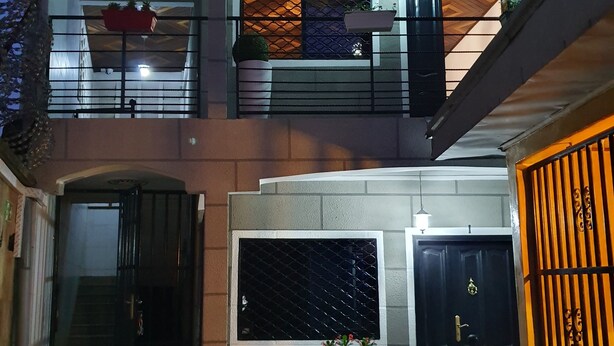 Um edifício de vários andares com entrada protegida por grades, uma escadaria que leva ao andar superior e plantas em vasos na varanda.