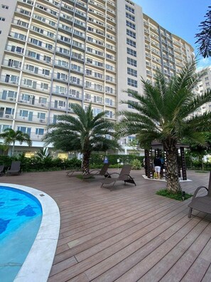 Un hotel con piscina, sillas de descanso, palmeras y un edificio grande al fondo.