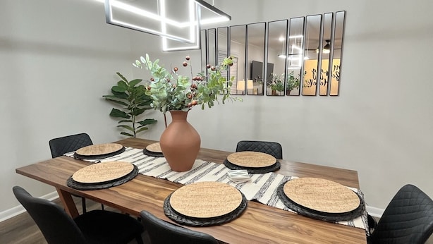 Ein moderner Essbereich mit Holztisch, schwarzen Stühlen, gewebten Tischsets, einer Vase mit Blumen und einer geometrischen Leuchte.