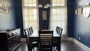 A dining room with a dark wood table and chairs, two windows with sheer curtains, a chandelier, and a dresser with drawers.
