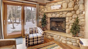 Una sala de estar acogedora con chimenea de piedra, un sofá de cuadros y vistas a un paisaje nevado a través de puertas de cristal amplias.