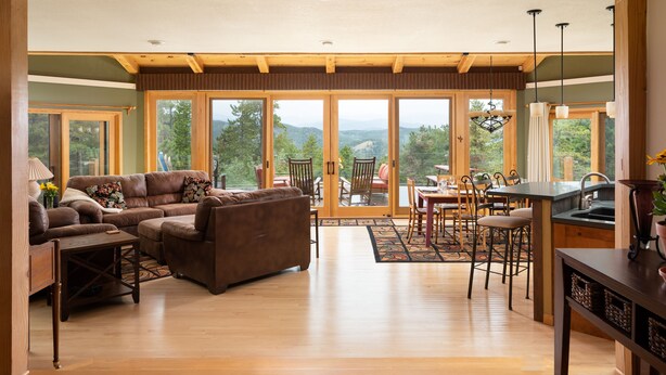 A spacious living area with a large sofa, dining table, and chairs, wooden flooring, and large windows offering a view of the outdoors. - Spacious 4 BR mountain home with pool table, hot tub, and incredible views (Golden)