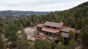 A modern two-story house with a deck, surrounded by dense forest and mountains in the background. - Spacious 4 BR mountain home with pool table, hot tub, and incredible views (Golden)