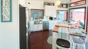 Una cocina compacta con armarios blancos, un refrigerador de acero inoxidable, un microondas y una isla de madera con un lavamanos. Hay lámparas colgantes sobre la isla y una ventana con vista a un patio trasero.