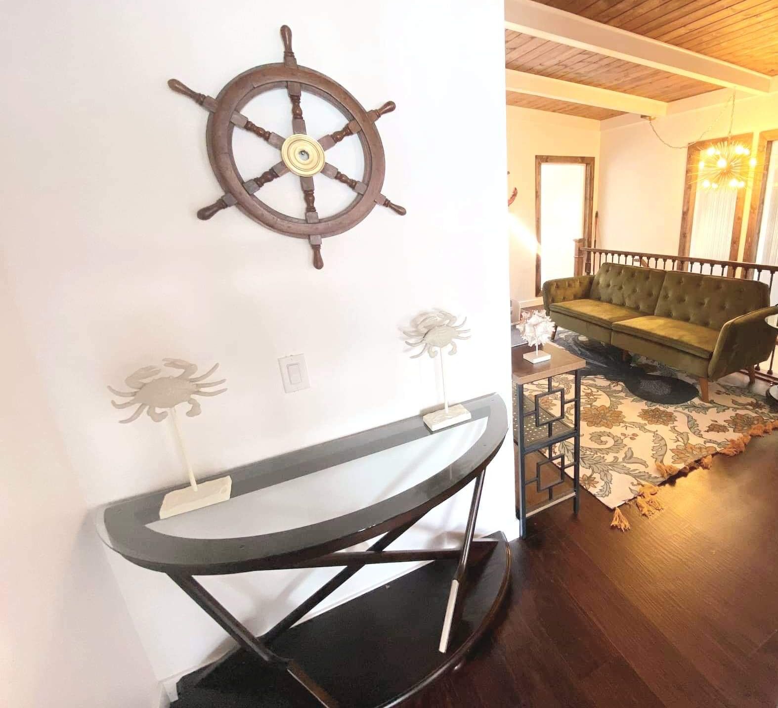 A room with a wooden ship's wheel on the wall, a grey console table with a white flower sculpture, and a green sofa with a patterned rug.