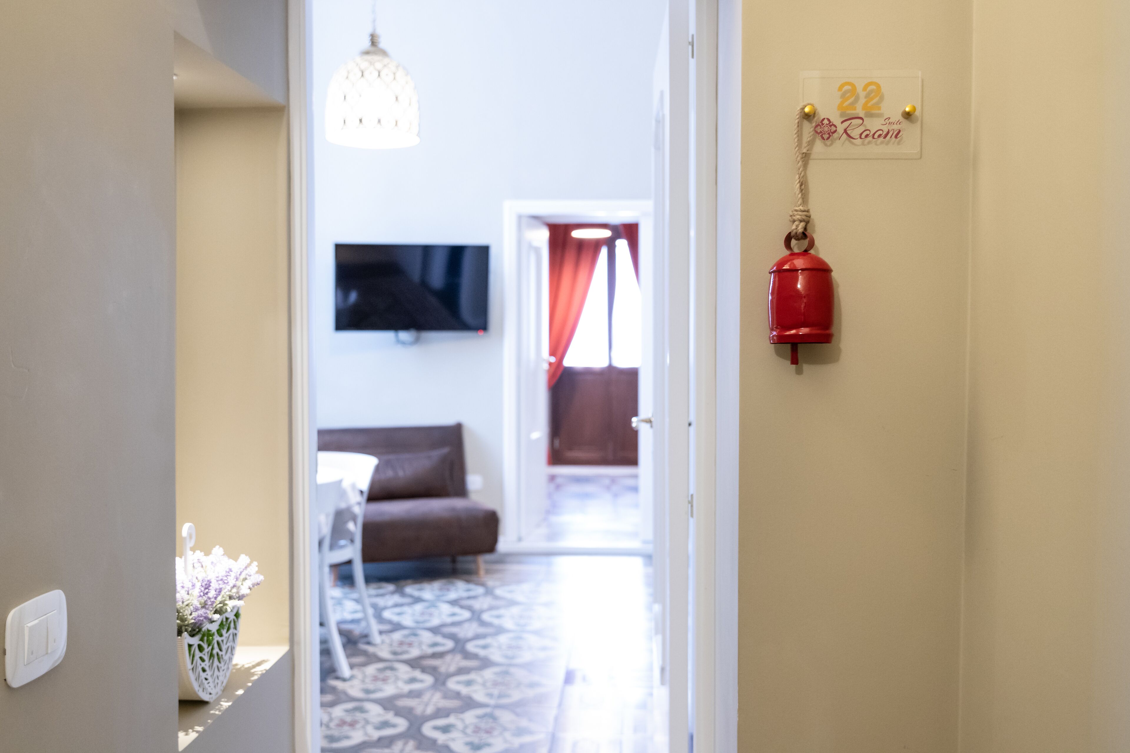 Un corridoio che conduce a un soggiorno con divano, televisore a parete, pavimento decorato e una campanella rossa.