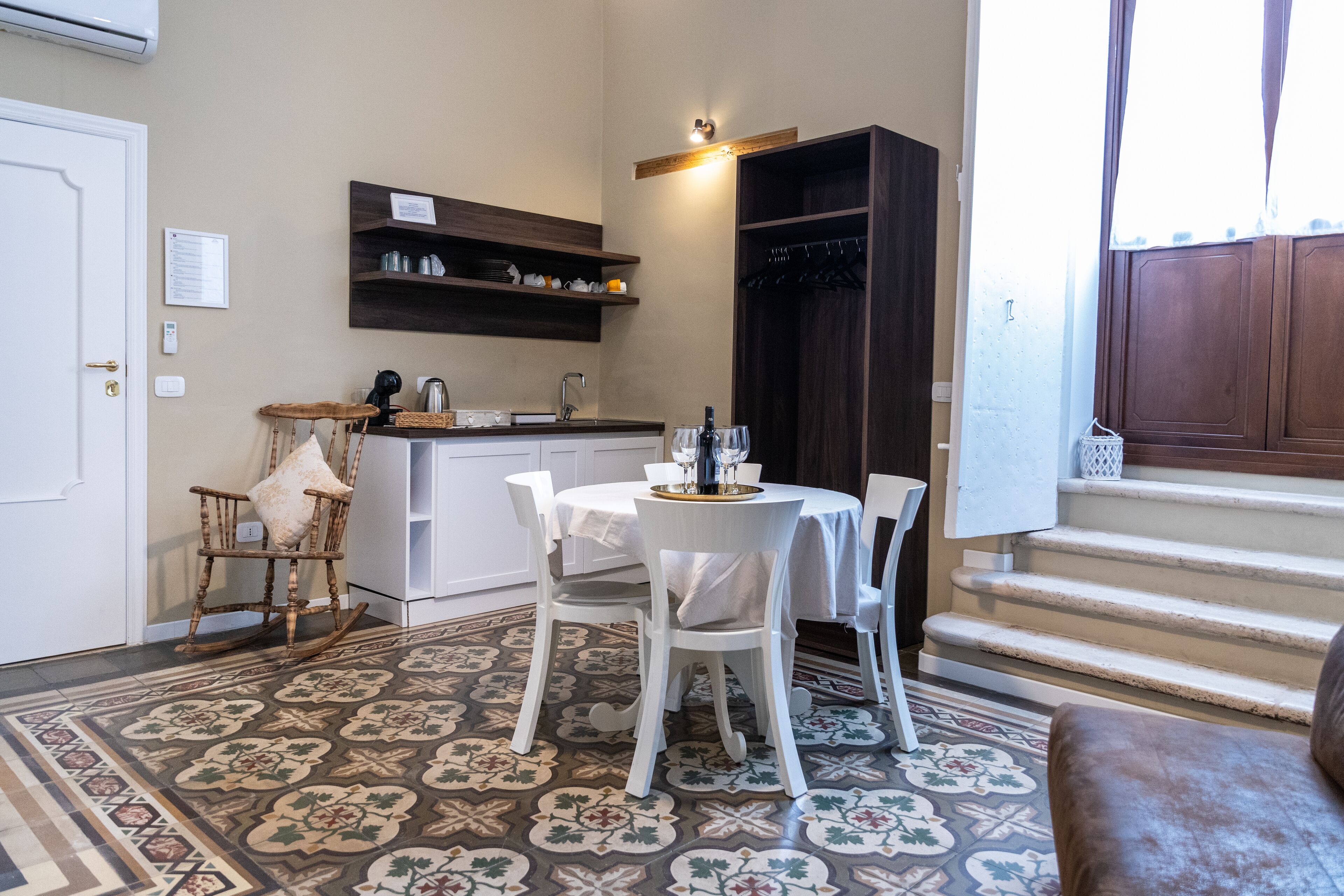 A compact kitchen with white cabinetry, a dining area with a round table and chairs, a patterned floor, and a staircase leading to a dark wooden door.