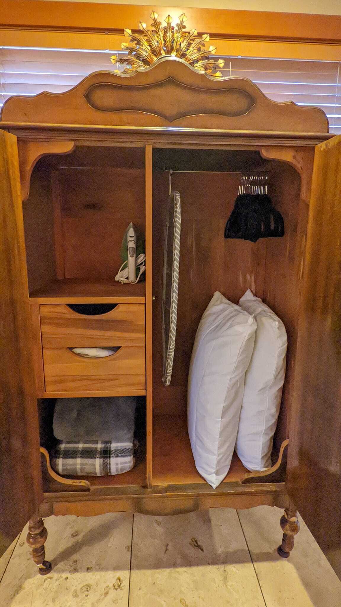 A wooden wardrobe with a mirror, a hanging rack, and a bed with white bedding.