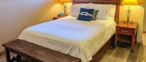 A neatly made bed with a wooden headboard, two bedside lamps, a bench, and a framed picture on the wall.