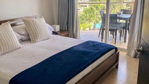 Una camera d'hotel con un letto, un comodino, una finestra vista piscina e edifici, e un balcone con sedie e un tavolo.