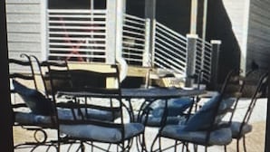 Uma área de estar coberta ao ar livre, com cadeiras e mesa de metal, guarda-corpo branco e um edifício ao fundo.