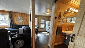 A cozy cabin interior with a wooden floor, a sofa, a dining table, and a kitchen area with a sink and cabinets. - COZY BEAR LODGE-On UTV/Snowmobile trail, Beachside Cabin w/ Porch, Private dock (St. Germain)