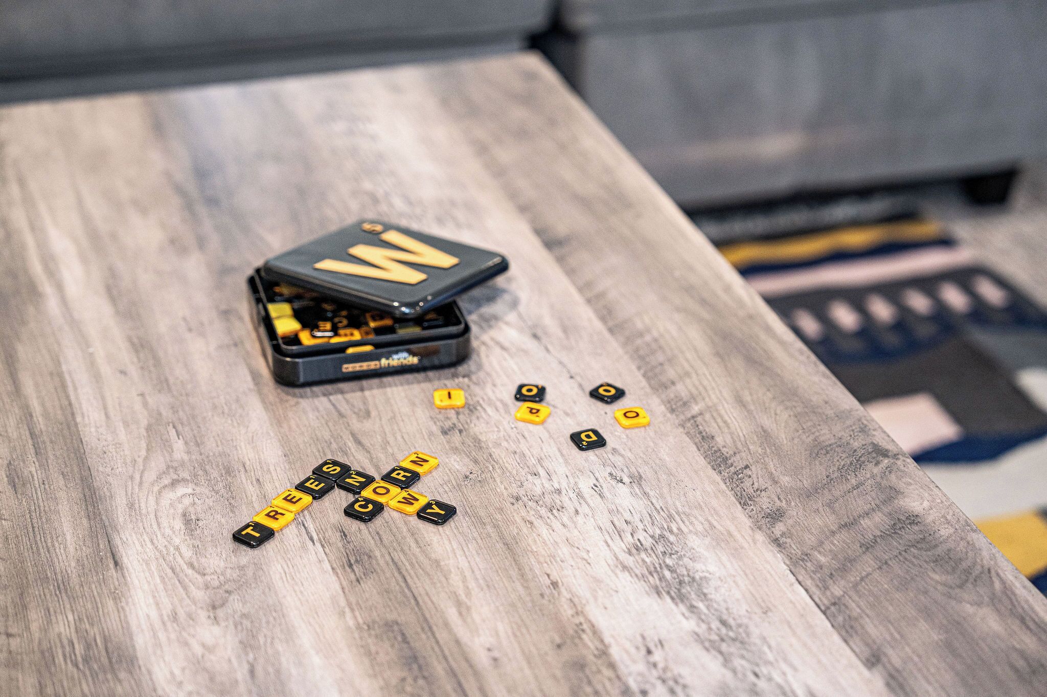 A wooden table with a Scrabble game in progress, featuring tiles spelling out words like "TREE" and "CORN."