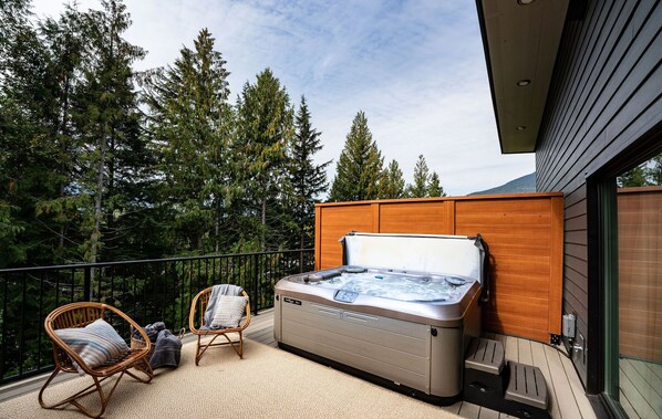 Ein Whirlpool auf einem Balkon mit einem Holzkopfteil, umgeben von einem Korbstuhl und einem Rattan-Sessel, mit Blick auf hohe Bäume.