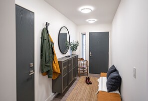 Ein Flur mit Kleiderständer, Spiegel, Topfpflanze, Stuhl und einem Bett mit Kissen.