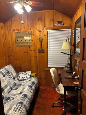 Una habitación acogedora de estilo cabaña con paredes de madera, un sofá de cuadros, un escritorio con lámpara y una ventana con persianas.