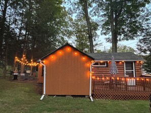 Una cabaña de madera con techo inclinado, rodeada por una terraza de madera y luces colgantes.