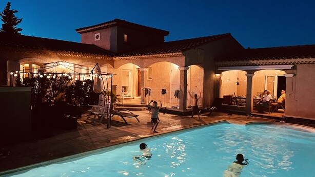 Ein Swimmingpool mit zwei Personen im Wasser, umgeben von einer überdachten Terrasse mit Sitzgelegenheiten und Lichterketten.