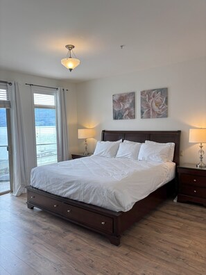 Une chambre à coucher avec un grand lit, deux tables de chevet avec des lampes, un luminaire au plafond et une vue sur la mer par la fenêtre.