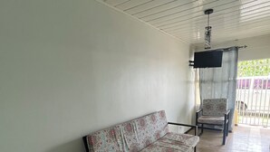 Ein Zimmer mit einer Couch und einem Sessel, einem an der Wand befestigten Fernseher und einem Fenster mit Blick auf einen Balkon.