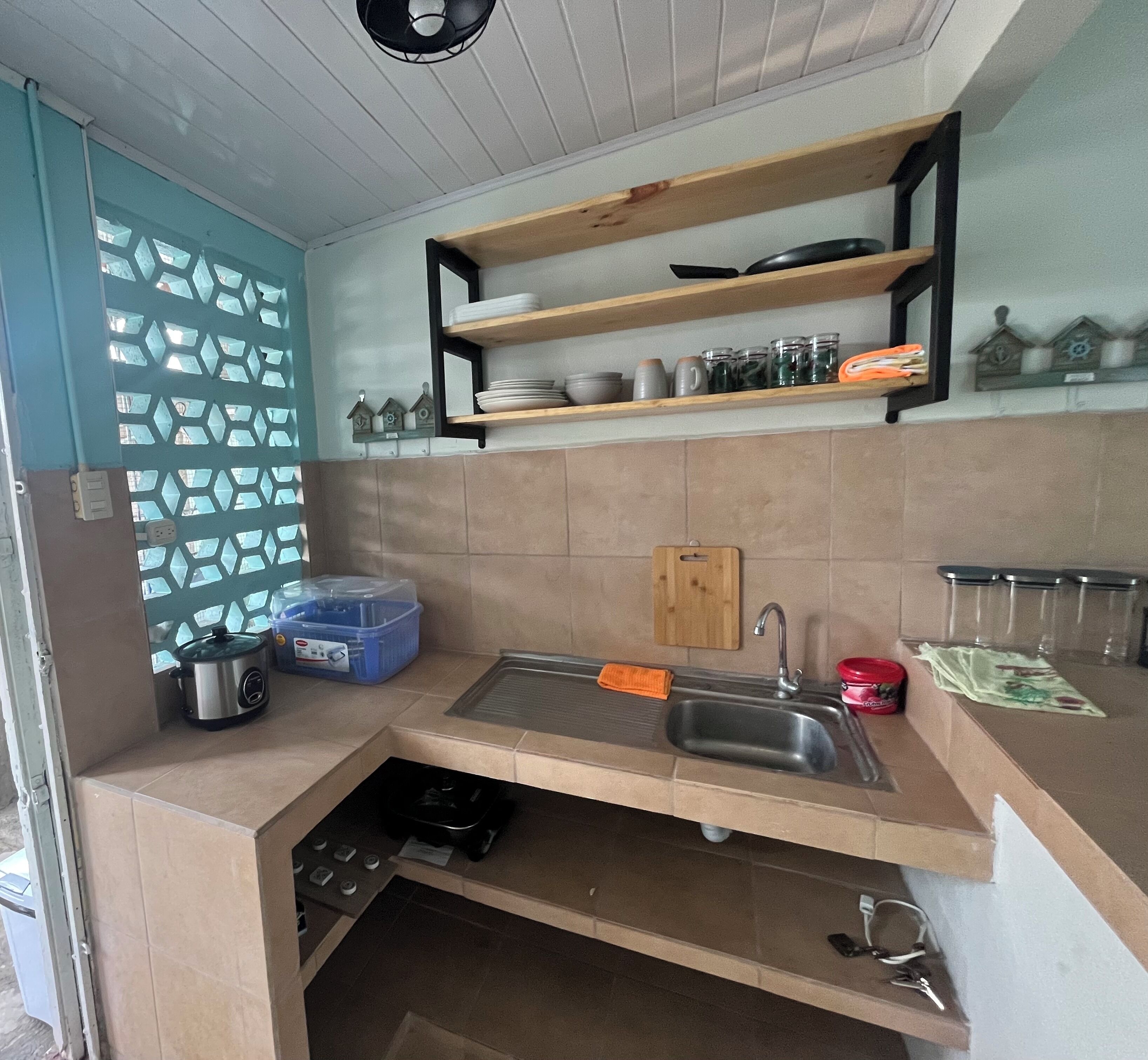 Cocina compacta con lavamanos, tabla de cortar y repisas superiores con platos y frascos.