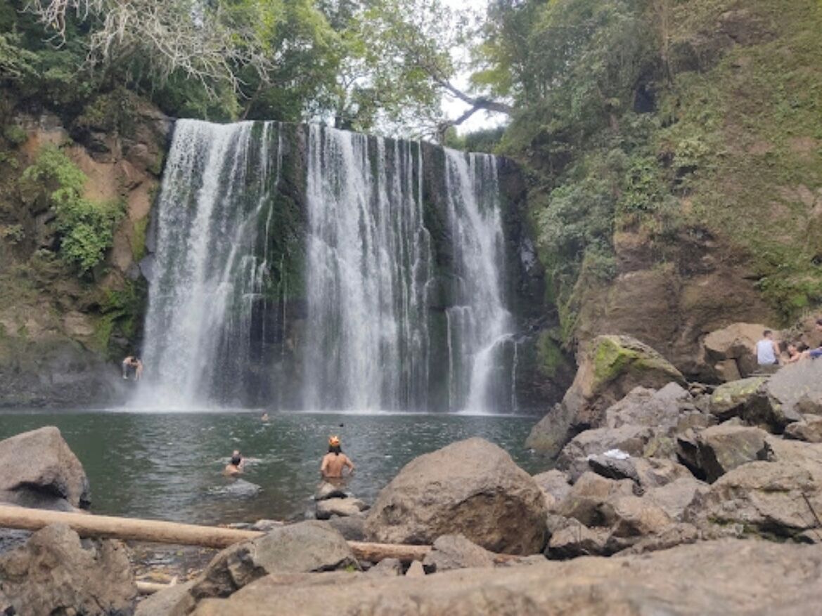 Una cascada con múltiples corrientes que caen en una poza rodeada de rocas y follaje.