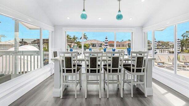 A bright dining area with white chairs and a table, large windows, and a view of a resort. -  Luxury WaterFront Home, Putting Green, Bocce, Pool/PPong Tbl (Marco Island)