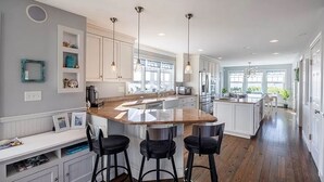 Una cocina moderna con una isla central, taburetes de bar, iluminación colgante y vista al comedor.
