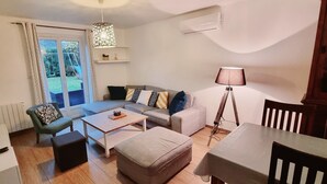 Un salon comprenant un canapé, une table basse et une table à manger avec des chaises. On y trouve une fenêtre avec des rideaux, un climatiseur et une lampe.